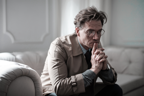 Distressed man resting his chin on his folded hands internally debating while sitting on a couch.